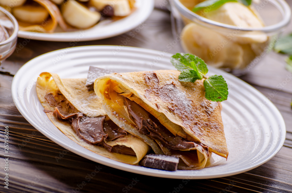 French crepes with chocolate sauce and banana in ceramic dish on wooden kitchen table French crepes with chocolate sauce and banana in ceramic dish on wooden kitchen table