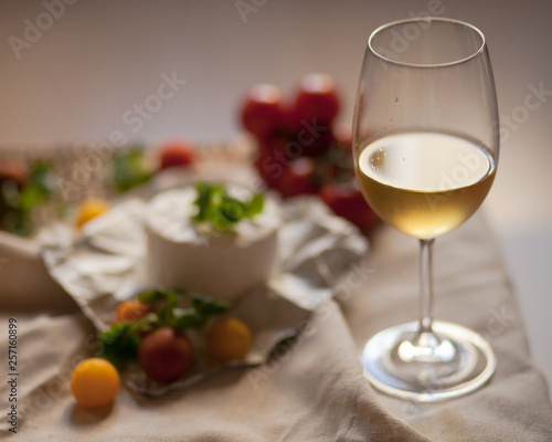 Glass of white wine, tasty starter of french cheese served with green salad and tomatoes on rustic cloth. Evening light, dark image