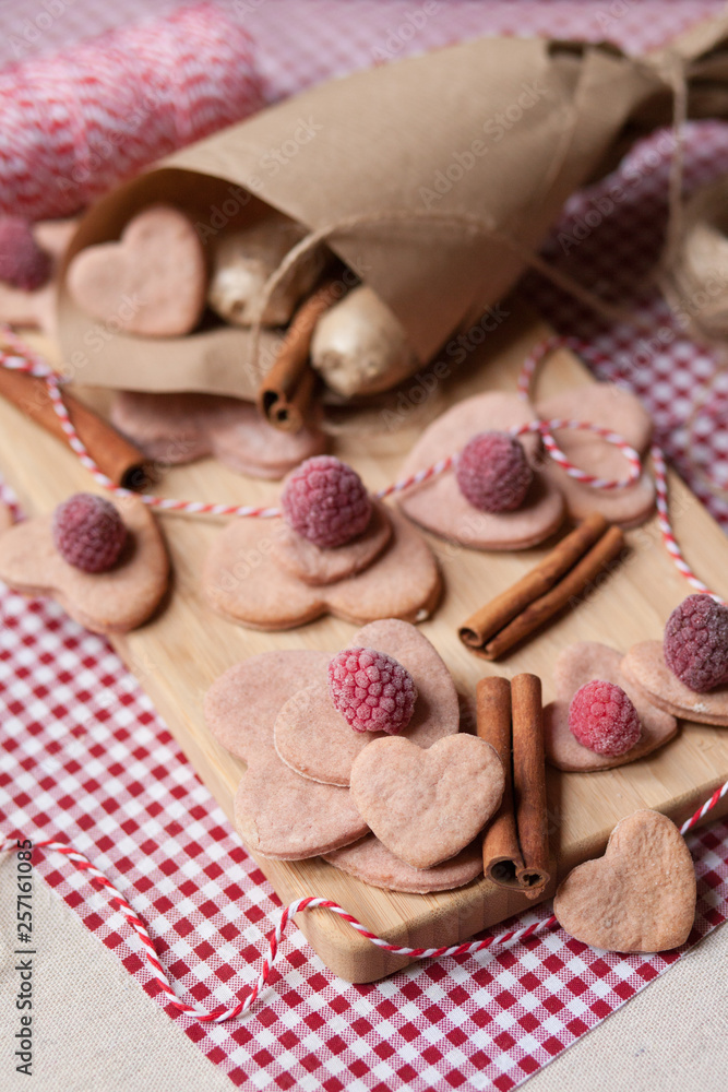 Hand made Saint Valentine's day surprise: home made pink sweet cookies in the shape of heart with a frozen raspberries served on a wooden boar and red napkin. Craft paper cornet on a background