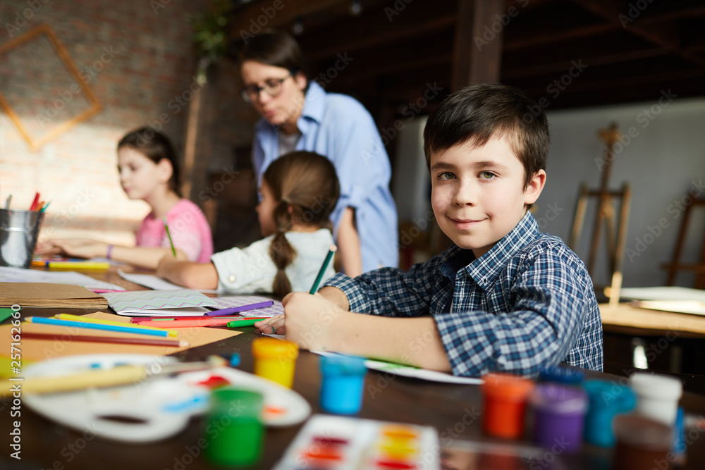 Fototapeta premium Portrait of cute little boy looking at camera while painting pictures in art class, copy space