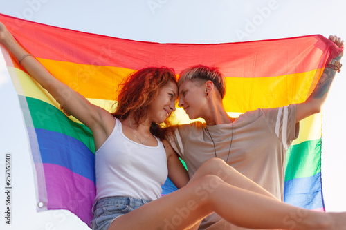Fotografie, Obraz Beautiful lesbian young couple gently lovingly hugging with rainbow flag, equal