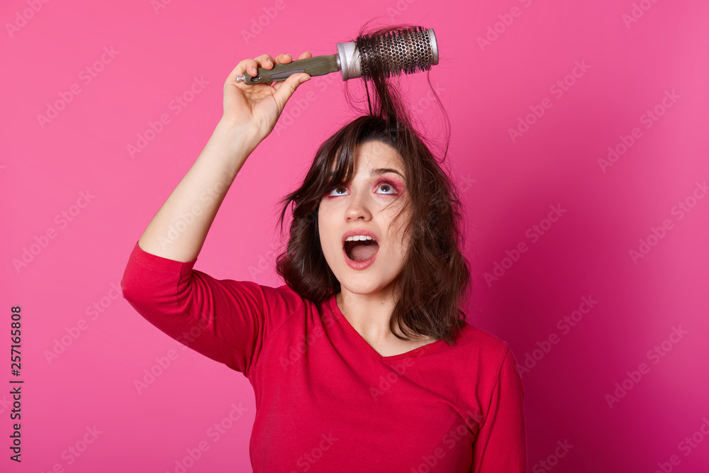 Adorable woman with brown tangled hair tries to comb hairs, looks up, wants to blow out comb and doing new hairstyle, dressesd in casual jumper, poses against pink wall, copy space for advertisment. Adorable woman with brown tangled hair tries to comb hairs, looks up, wants to blow out comb and doing new hairstyle, dressesd in casual jumper, poses against pink wall, copy space for advertisment.