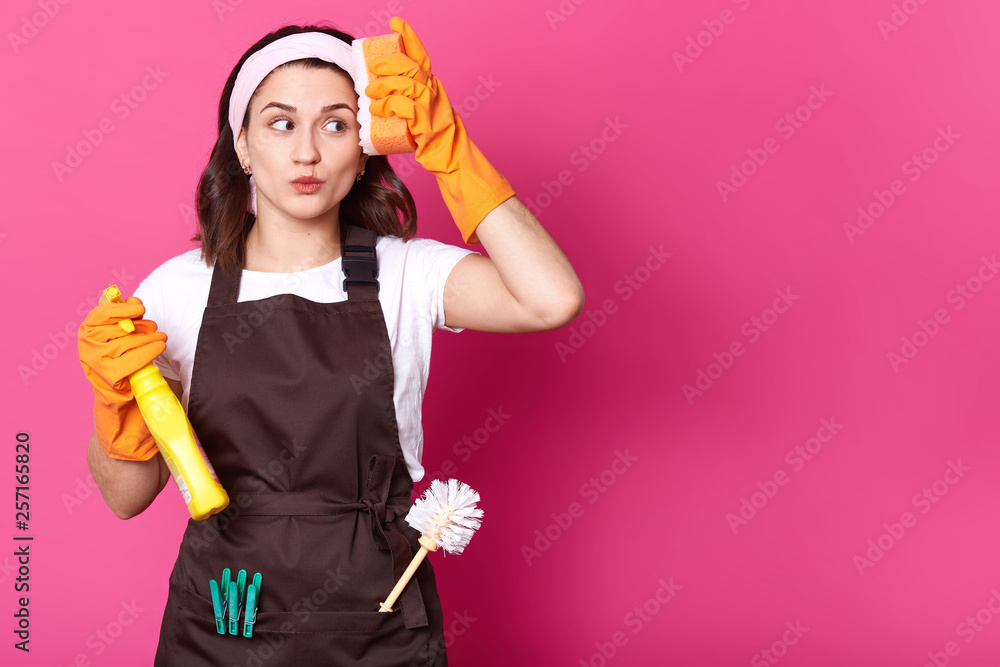 Portrait of happy young woman with orange sponge and detergent in hands, cleans her house wearing orange rubber gloves for hands protection and brown apron, isolated over pink background. Copy space. Portrait of happy young woman with orange sponge and detergent in hands, cleans her house wearing orange rubber gloves for hands protection and brown apron, isolated over pink background. Copy space.