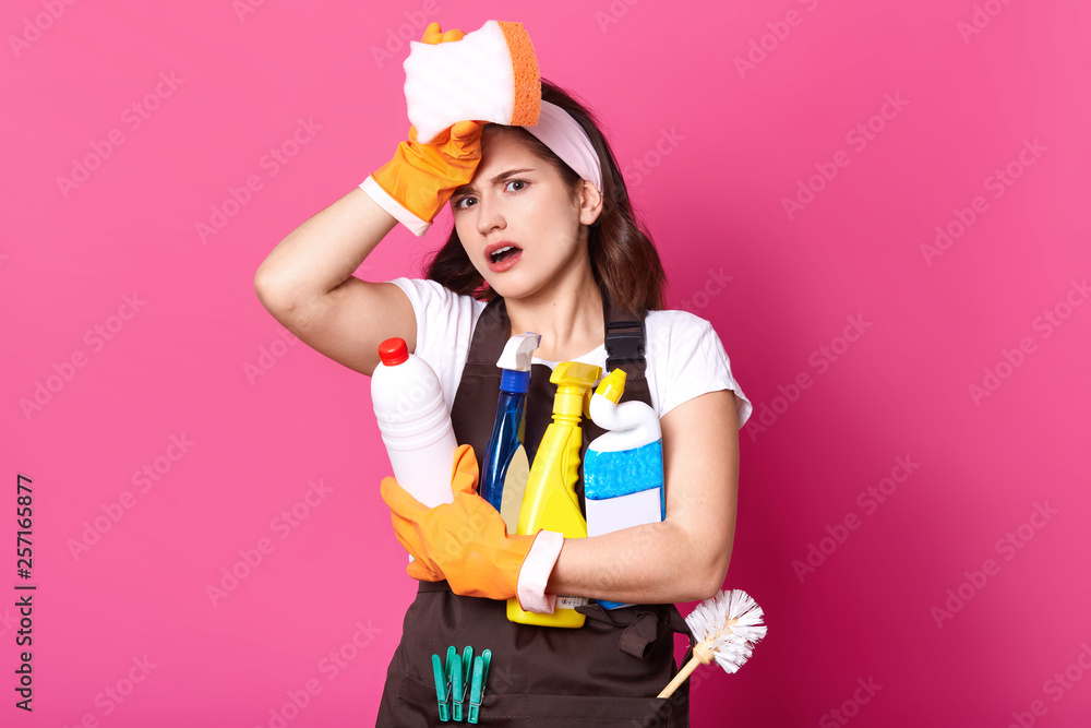 Young tired housewife in brown apron and white casual t shirt, with cleaning deretgents, isolated on pink background. Female has exuasted facial expressions, poses in photo sudio. Hygiene concept. Young tired housewife in brown apron and white casual t shirt, with cleaning deretgents, isolated on pink background. Female has exuasted facial expressions, poses in photo sudio. Hygiene concept.