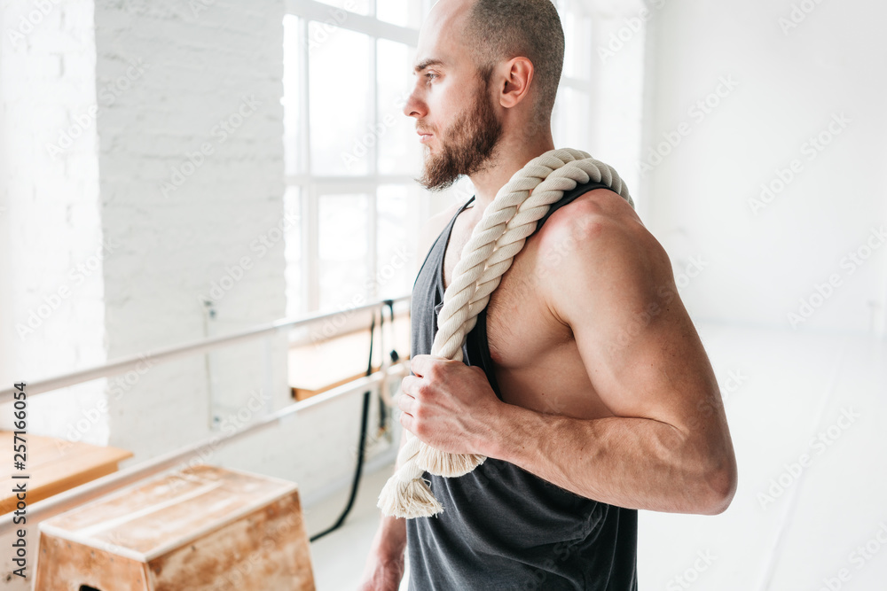 Portrait of fit man with battle ropes at cross gym. Strong male athlete holding on shoulder rope after intense workout Portrait of fit man with battle ropes at cross gym. Strong male athlete holding on shoulder rope after intense workout