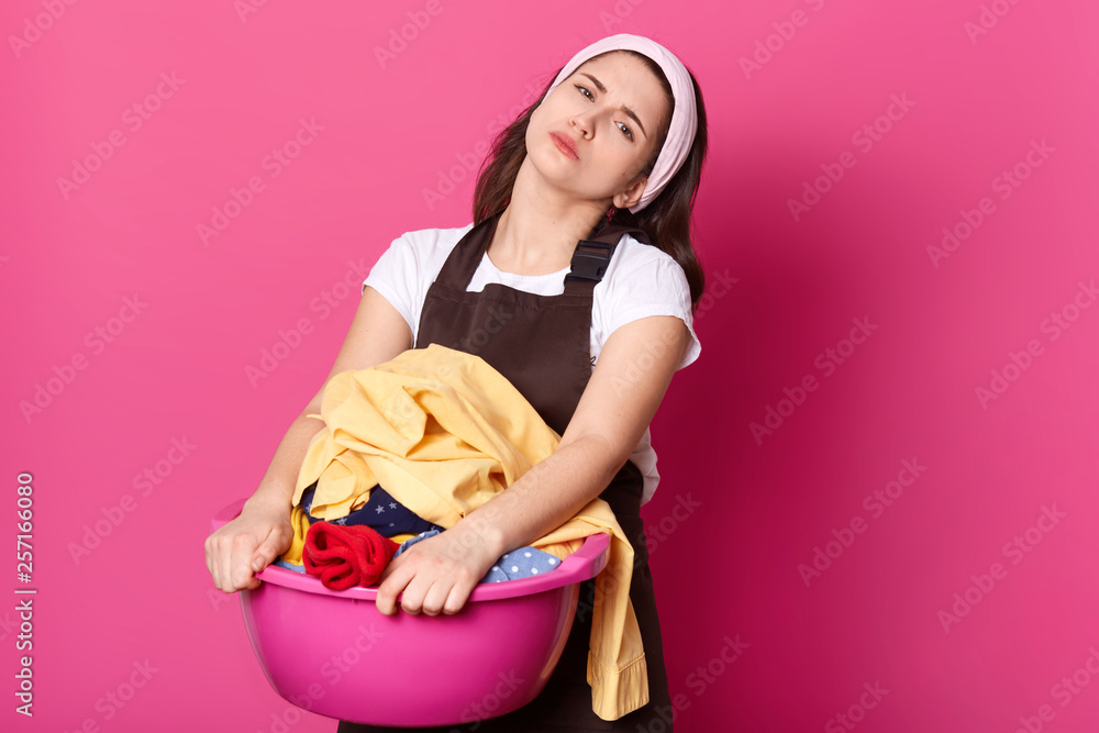 Image of upset European woman holds basin with dirty clothes, wears brown apron, has upset facial expressions, prepares for washing, does not want do household chores, poses on rosy studio background Image of upset European woman holds basin with dirty clothes, wears brown apron, has upset facial expressions, prepares for washing, does not want do household chores, poses on rosy studio background