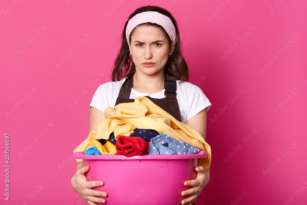 Portrait of caucasian housewife with white hair band and brown apron, holds basin of clothes, has upset facial expressions, does washing at home, beeing in bad mood, isolated over pink background. Portrait of caucasian housewife with white hair band and brown apron, holds basin of clothes, has upset facial expressions, does washing at home, beeing in bad mood, isolated over pink background.