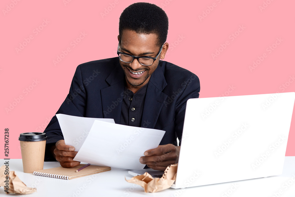 Portrait of cheerful male financier with dark skin, wears glasses, has fan expression, prepares report, drinks coffee, laughing while reads documents isolated over pink background. Business concept. Portrait of cheerful male financier with dark skin, wears glasses, has fan expression, prepares report, drinks coffee, laughing while reads documents isolated over pink background. Business concept.
