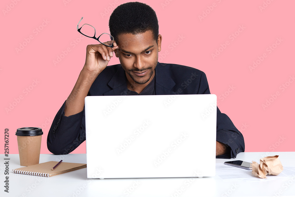 Young black business man with laptop, sits at white table, holds his spectacles in hand, has thoughtful facial expressions, thinks about important contract, poses isolated over rose background. Young black business man with laptop, sits at white table, holds his spectacles in hand, has thoughtful facial expressions, thinks about important contract, poses isolated over rose background.