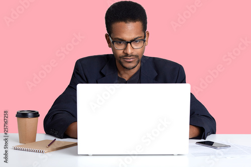 Close up portrait of dark skinned male wears black suit, works online with lap top, African American freelancer sits at white table in front of opened laptop computer, isolated over pink background.
