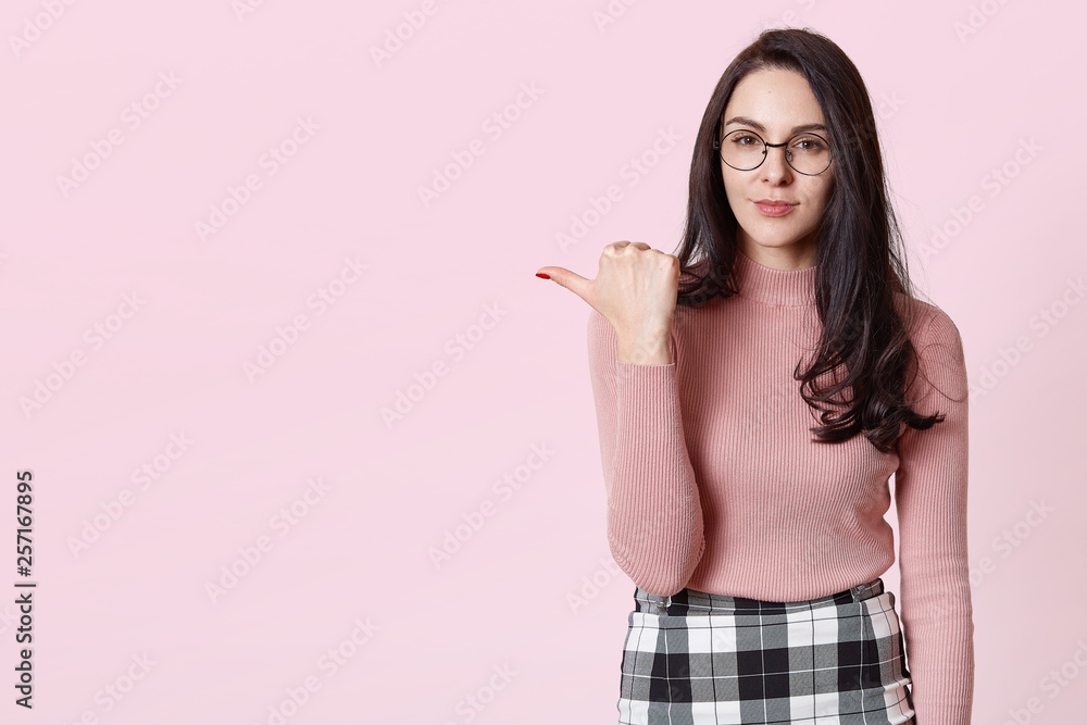 Portrait of stylish beautiful young woman isolated on rose background. Dark haired female points aside with her fore finger, shows copy space for your text. Attractive girl poses in photo studio. Portrait of stylish beautiful young woman isolated on rose background. Dark haired female points aside with her fore finger, shows copy space for your text. Attractive girl poses in photo studio.
