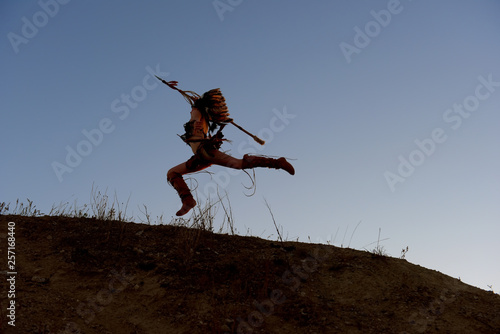 A young girl plays the part of a native American Indian girl. She poses on top of a hill wit the sun setting behind her.She leaps through the air in an attacking posture.