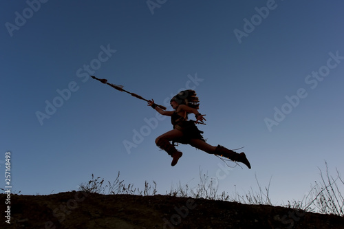 A young girl plays the part of a native American Indian  girl. She poses on top of a hill wit the sun setting behind her.She leaps through the air in an attacking posture.