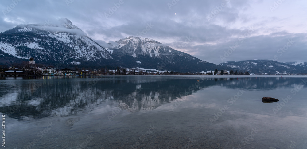 Panorama Wolfgangsee mit der Ortschaft Strobl und schneebedeckten Bergen im Hintergrund die sich im klaren Wasser spiegeln und Mond