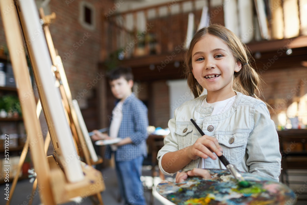 Waist up portrait of cute little girl painting picture on easel in art class and holding palette, copy space Waist up portrait of cute little girl painting picture on easel in art class and holding palette, copy space