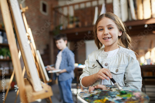 Waist up portrait of cute little girl painting picture on easel in art class and holding palette, copy space