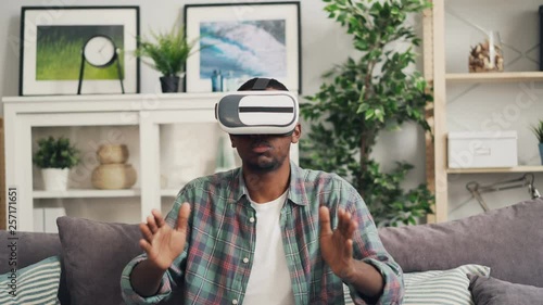 Zoom-in of African American young man wearing augmented reality glasses and moving hands smiling and emjoying game sitting on sofa in modern flat alone. photo