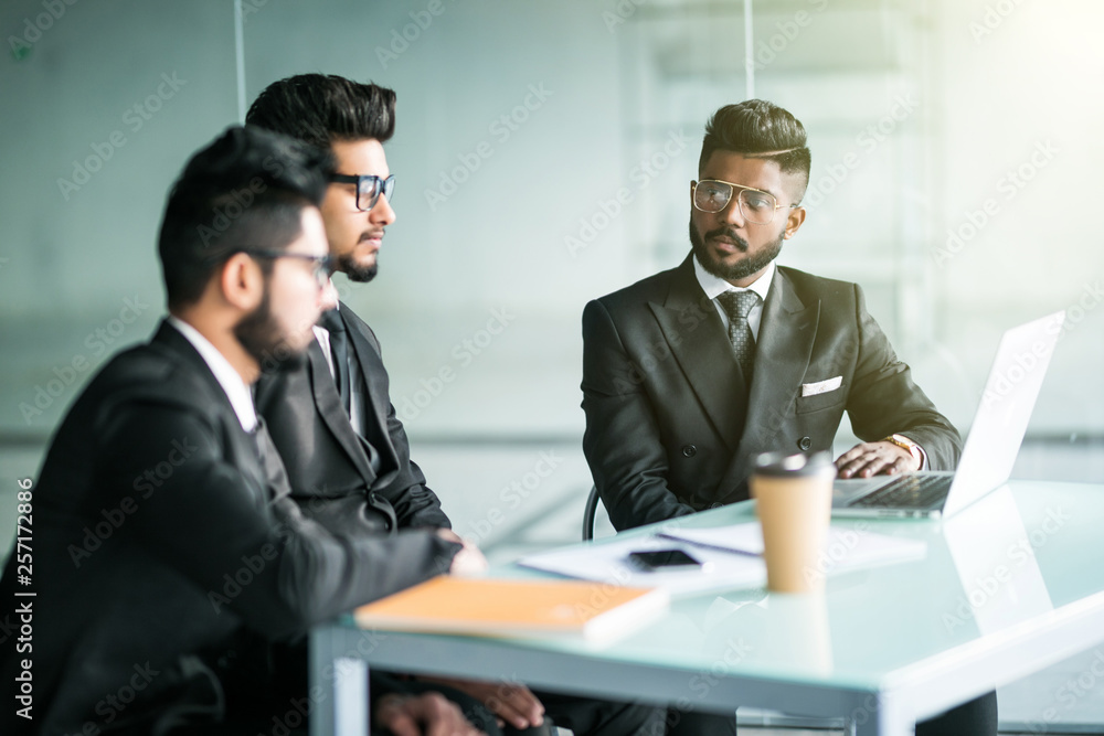 Three indian businessmen sitting around table meeting in modern open plan office Three indian businessmen sitting around table meeting in modern open plan office