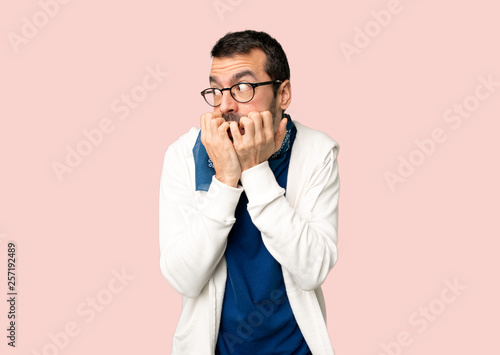Handsome man with glasses is a little bit nervous and scared putting hands to mouth on isolated pink background
