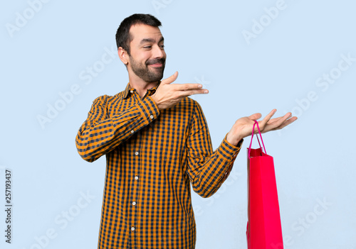 Man with shopping bags extending hands to the side for inviting to come on isolated blue background