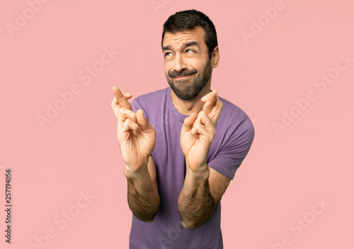 Handsome man with fingers crossing and wishing the best on isolated pink background