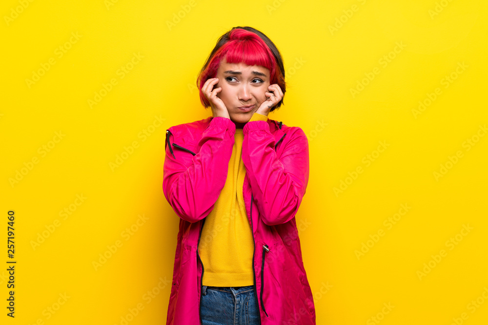 Young woman with pink hair over yellow wall frustrated and covering ears Young woman with pink hair over yellow wall frustrated and covering ears