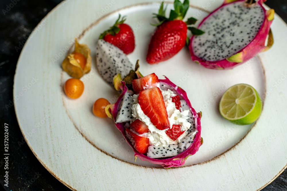 on a white, wooden plate, lie the fresh fruit of strawberries, among them a beautiful, pink dragon fruit filled with whipped cream and strawberries on a white, wooden plate, lie the fresh fruit of strawberries, among them a beautiful, pink dragon fruit filled with whipped cream and strawberries