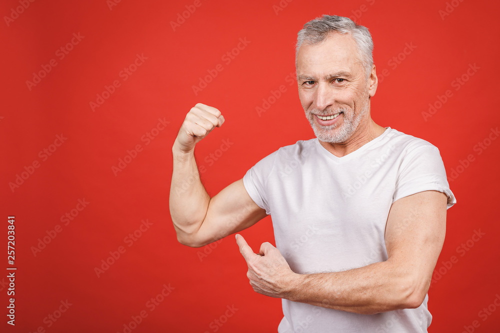 Check this out! Senior man showing muscle. Aged man flexing his arms isolated against red background. Cheerful excited modern cool pensioner grandpa practising bodybuilding. Check this out! Senior man showing muscle. Aged man flexing his arms isolated against red background. Cheerful excited modern cool pensioner grandpa practising bodybuilding.