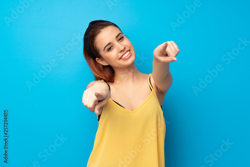 Young redhead woman over blue background points finger at you while smiling