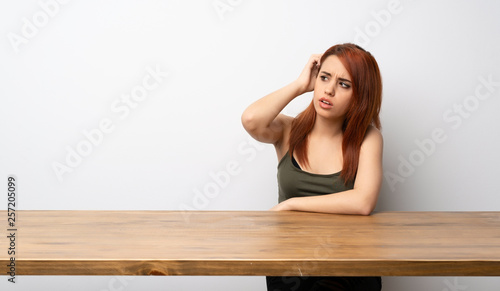 Young redhead woman at desk having doubts while scratching head