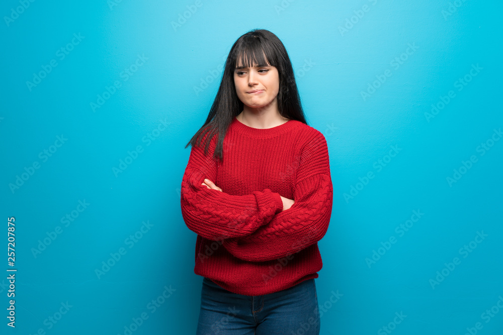 Woman with red sweater over blue wall with confuse face expression while bites lip Woman with red sweater over blue wall with confuse face expression while bites lip