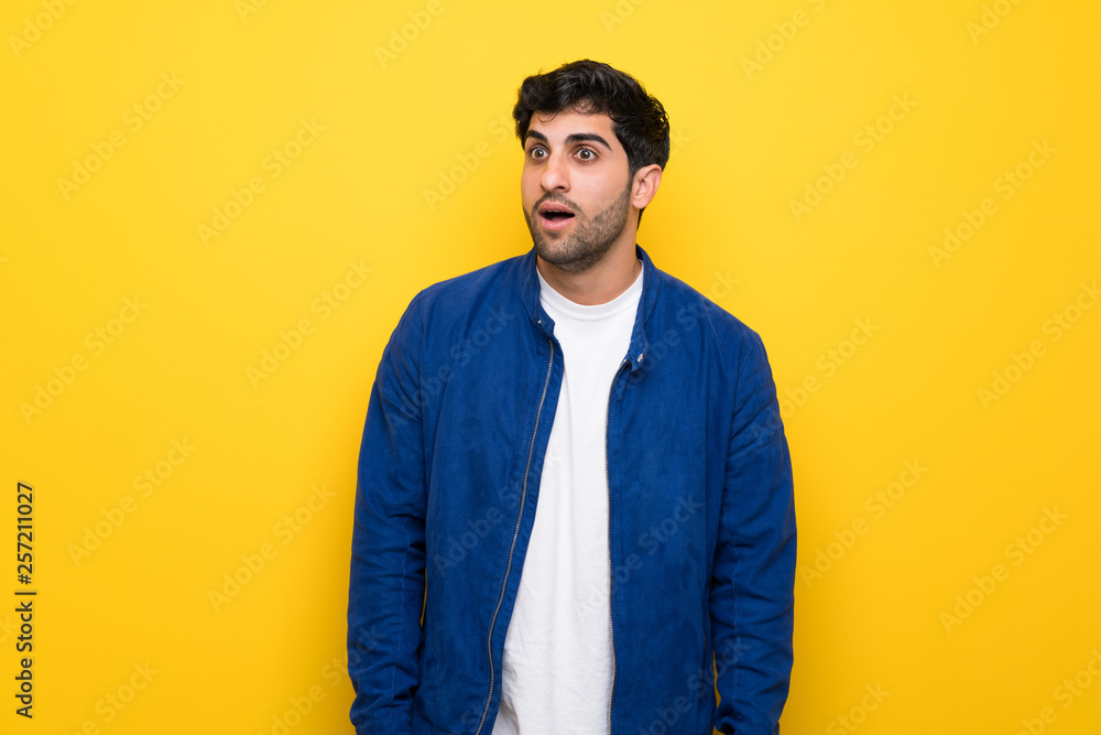Man with blue jacket over yellow wall with surprise facial expression Man with blue jacket over yellow wall with surprise facial expression