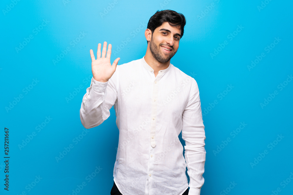 Young man over isolated blue wall saluting with hand with happy expression Young man over isolated blue wall saluting with hand with happy expression