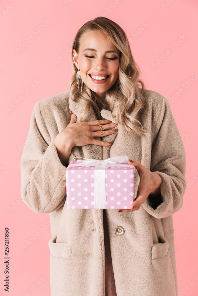 Pretty woman posing isolated over pink wall background dressed in fashion coat holding present box. Pretty woman posing isolated over pink wall background dressed in fashion coat holding present box.