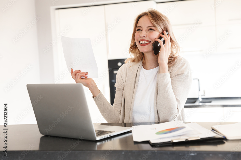 Beautiful blonde woman posing sitting indoors at home using laptop computer talking by mobile phone. Beautiful blonde woman posing sitting indoors at home using laptop computer talking by mobile phone.