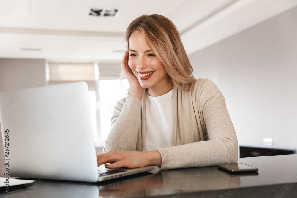 Beautiful blonde woman posing sitting indoors at home using laptop computer. Beautiful blonde woman posing sitting indoors at home using laptop computer.