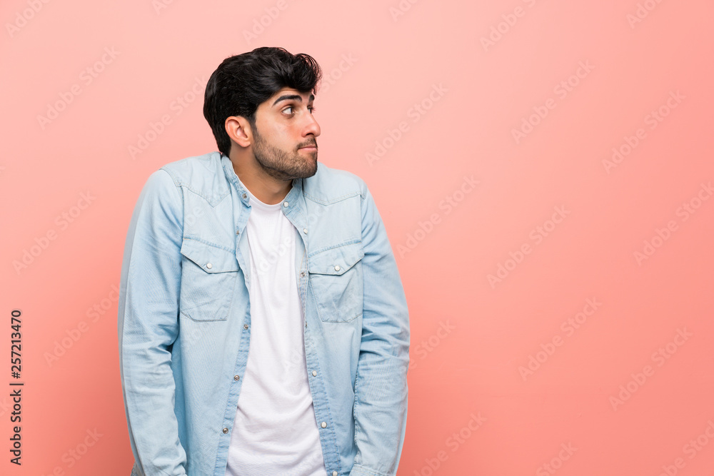Young man over pink wall making doubts gesture looking side Young man over pink wall making doubts gesture looking side