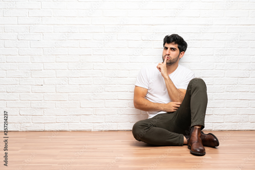 Young man sitting on the floor showing a sign of silence gesture putting finger in mouth Young man sitting on the floor showing a sign of silence gesture putting finger in mouth