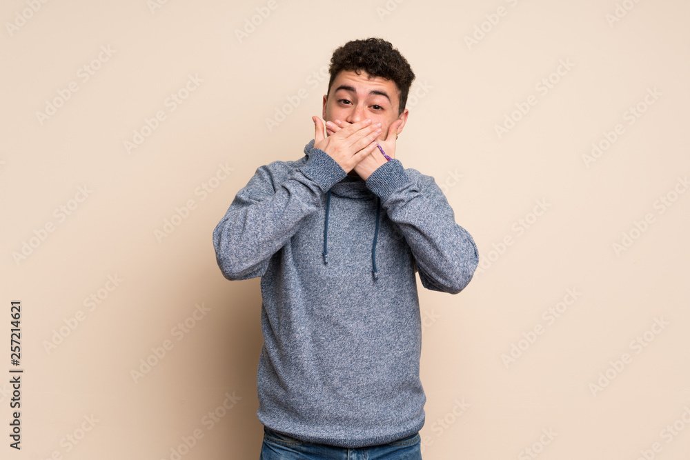 Young man over isolated wall covering mouth with hands Young man over isolated wall covering mouth with hands