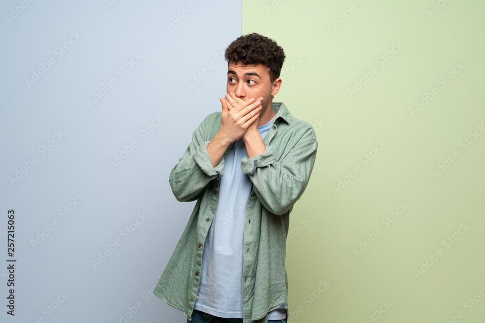Young man over blue and green background covering mouth with hands