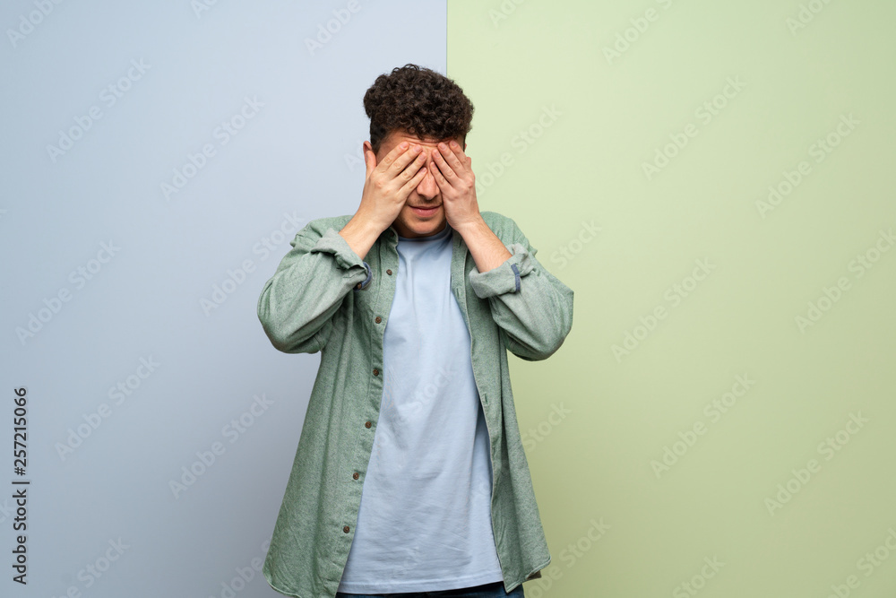 Young man over blue and green background covering eyes by hands. Surprised to see what is ahead Young man over blue and green background covering eyes by hands. Surprised to see what is ahead