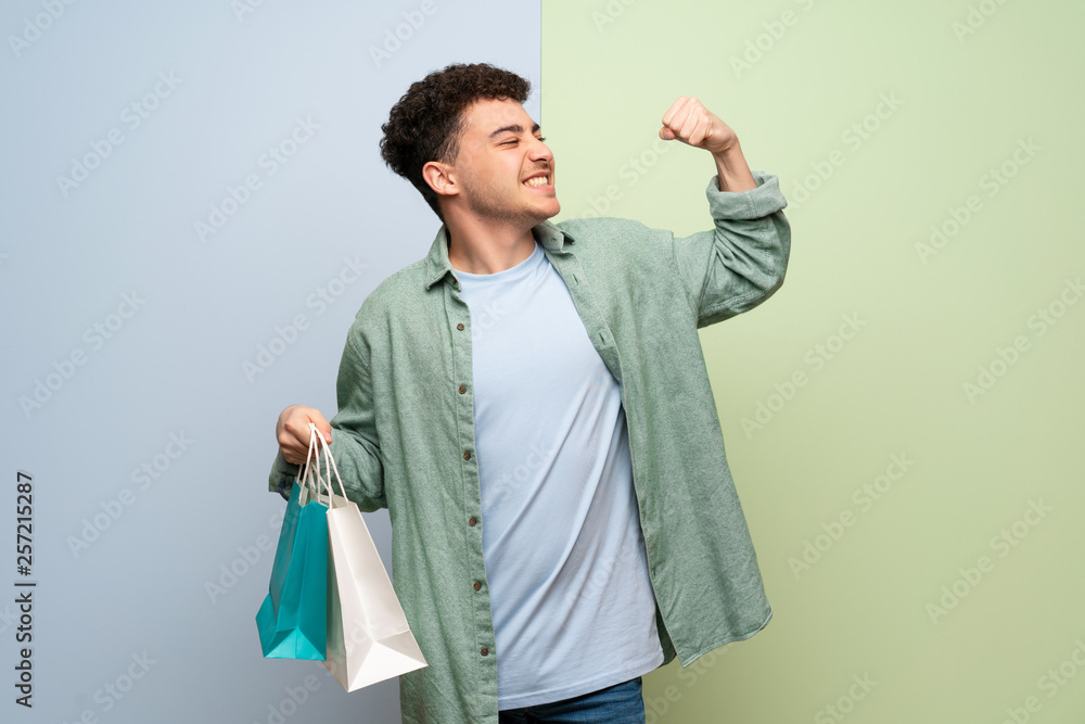 Young man over blue and green background holding a lot of shopping bags in victory position Young man over blue and green background holding a lot of shopping bags in victory position