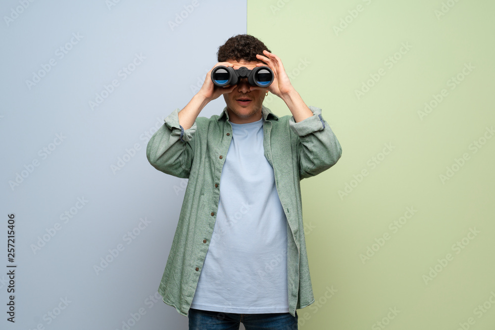 Young man over blue and green background and looking in the distance with binoculars Young man over blue and green background and looking in the distance with binoculars