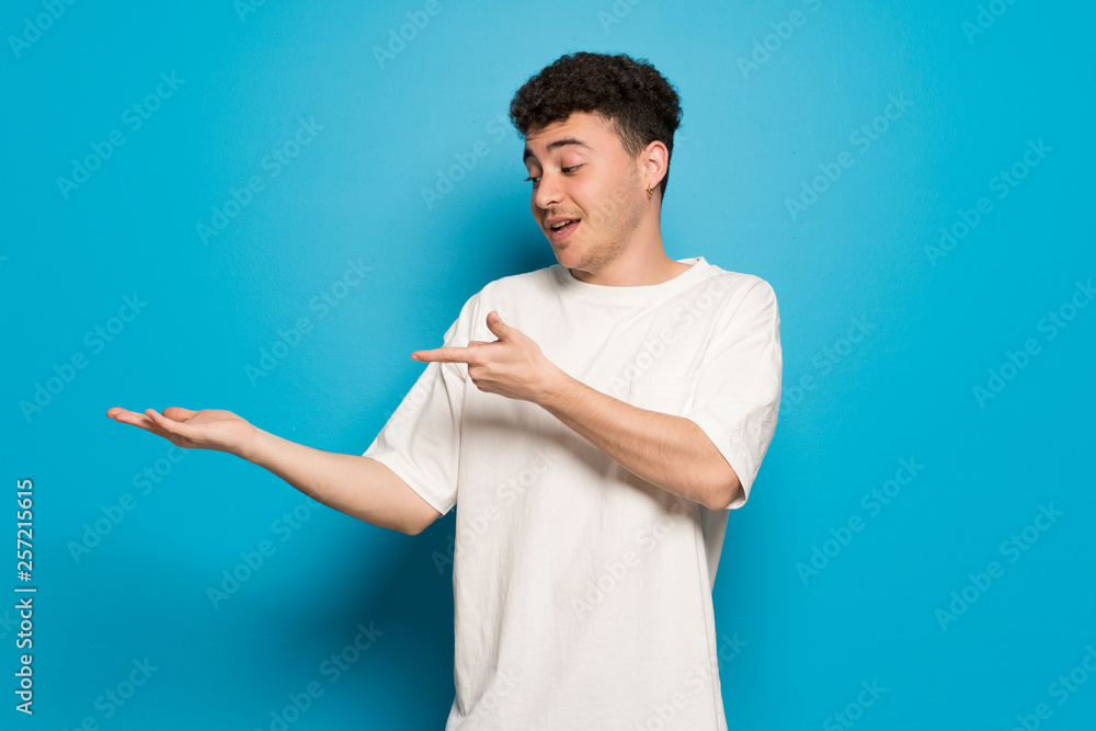 Young man over blue background holding copyspace imaginary on the palm to insert an ad