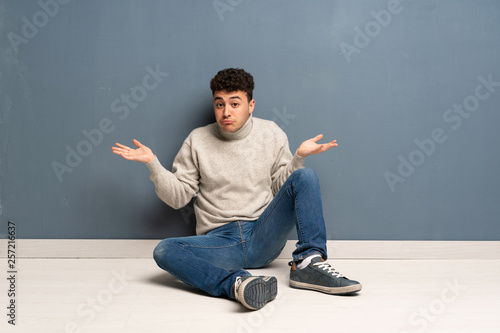 Young man sitting on the floor having doubts while raising hands