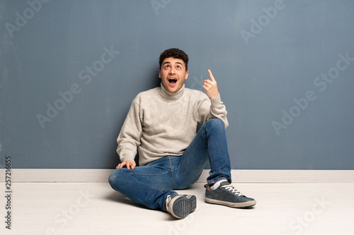 Young man sitting on the floor pointing up and surprised