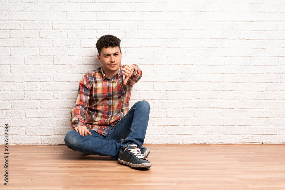 Young man sitting on the floor showing thumb down with negative expression Young man sitting on the floor showing thumb down with negative expression