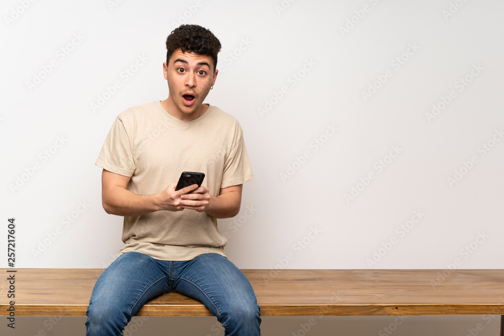 Young man sitting on table surprised and sending a message