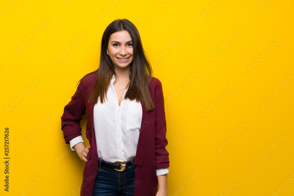 Young woman over yellow wall posing with arms at hip and smiling Young woman over yellow wall posing with arms at hip and smiling