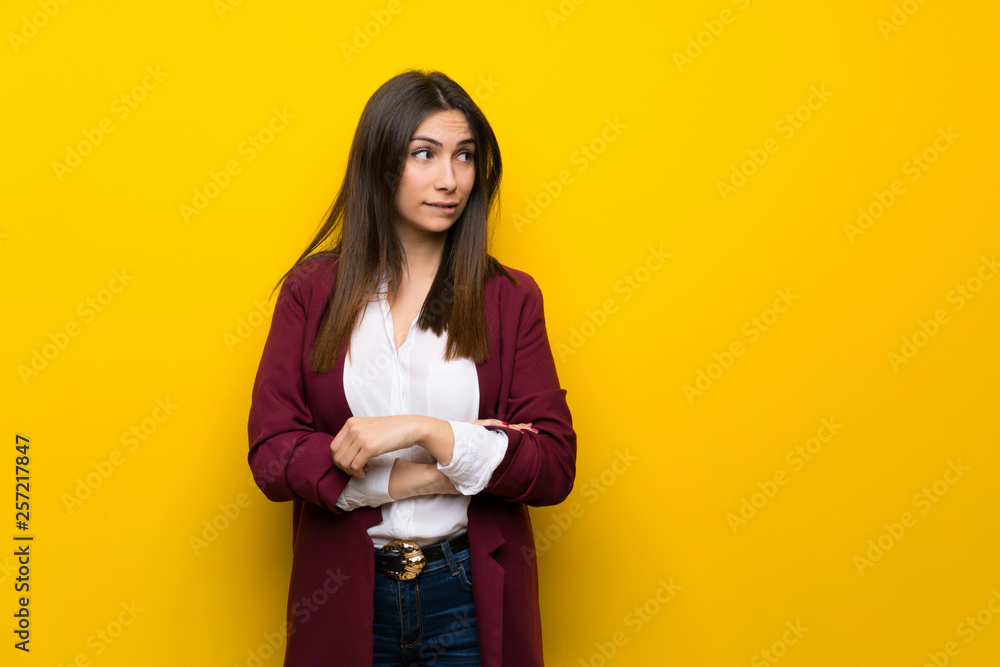 Young woman over yellow wall with confuse face expression while bites lip Young woman over yellow wall with confuse face expression while bites lip